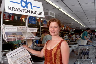846317 Afbeelding van een jonge vrouw die de krant Het Utrechts Nieuws pakt uit het krantenrek van supermarkt Albert ...